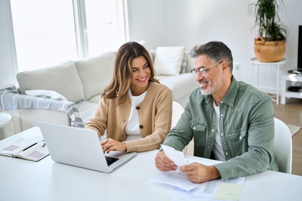 Happy middle aged couple sitting at a desk in their beautiful home reviewing their finances on a laptop.