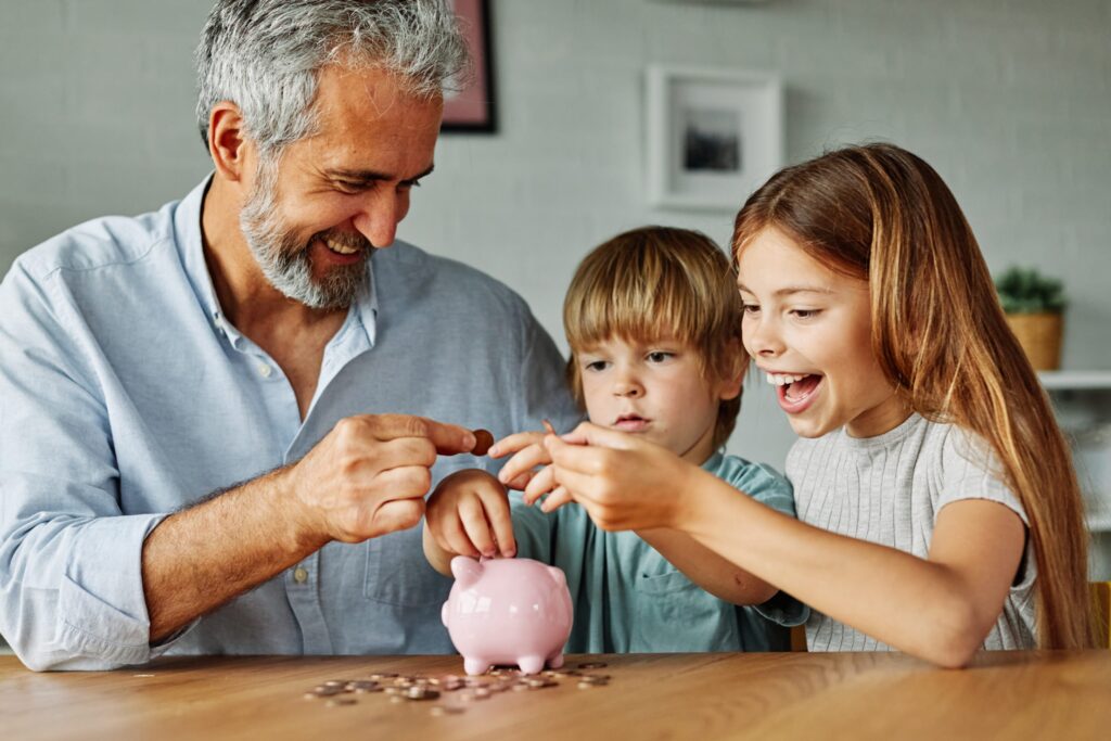 A middle aged man sat at a table with two young children. They have coins in their hands and are putting the money into a piggy bank.